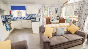 Een gezellige woonkamer met een bruine bank en fauteuil, een eethoek met tafel en stoelen, en een keuken met witte kasten en blauwe tegels.