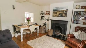 A living room with a fireplace, a dining area with a table and chairs, a bookshelf, and a large rug. - Rose Cottage (Bakewell)