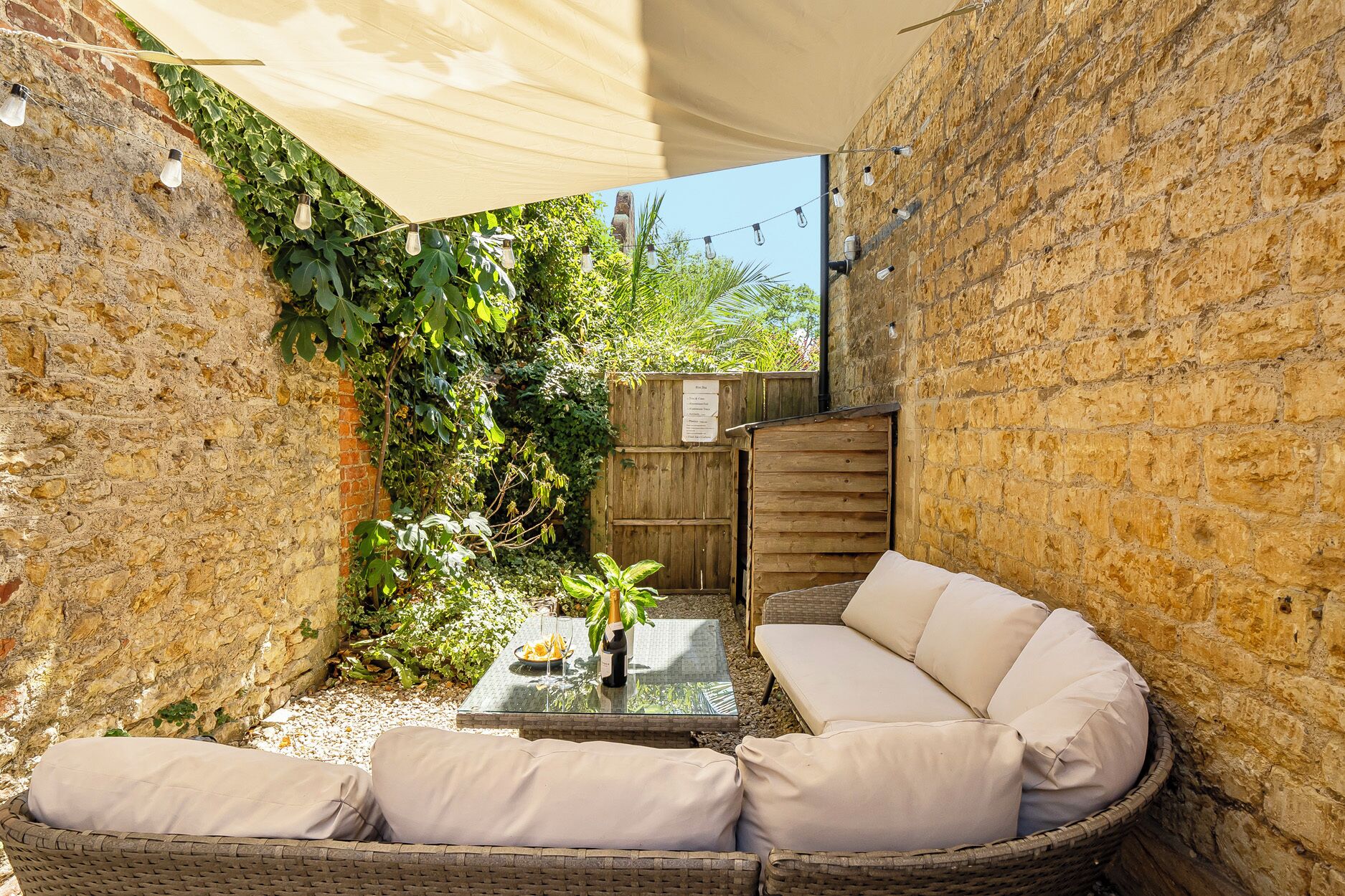 A cozy outdoor seating area with wicker furniture and a glass table, surrounded by stone walls and greenery.