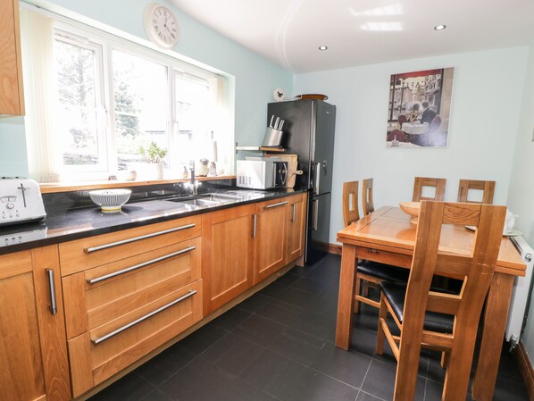 A kitchen with wooden cabinets, a black countertop, a refrigerator, a microwave, and a dining table with chairs. - 6 Stad Yr Ysgol (Blaenau Ffestiniog)