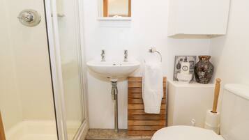 A bathroom with a glass shower enclosure, a white sink with two faucets, a wooden vanity, a toilet, and a small shelf with decorative items.