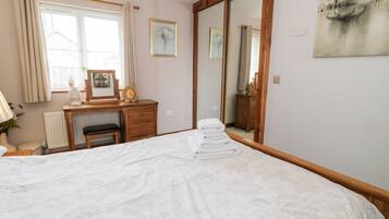 A neatly arranged bedroom with a bed, a wooden desk, a chair, a radiator, a window with curtains, and a large mirror.