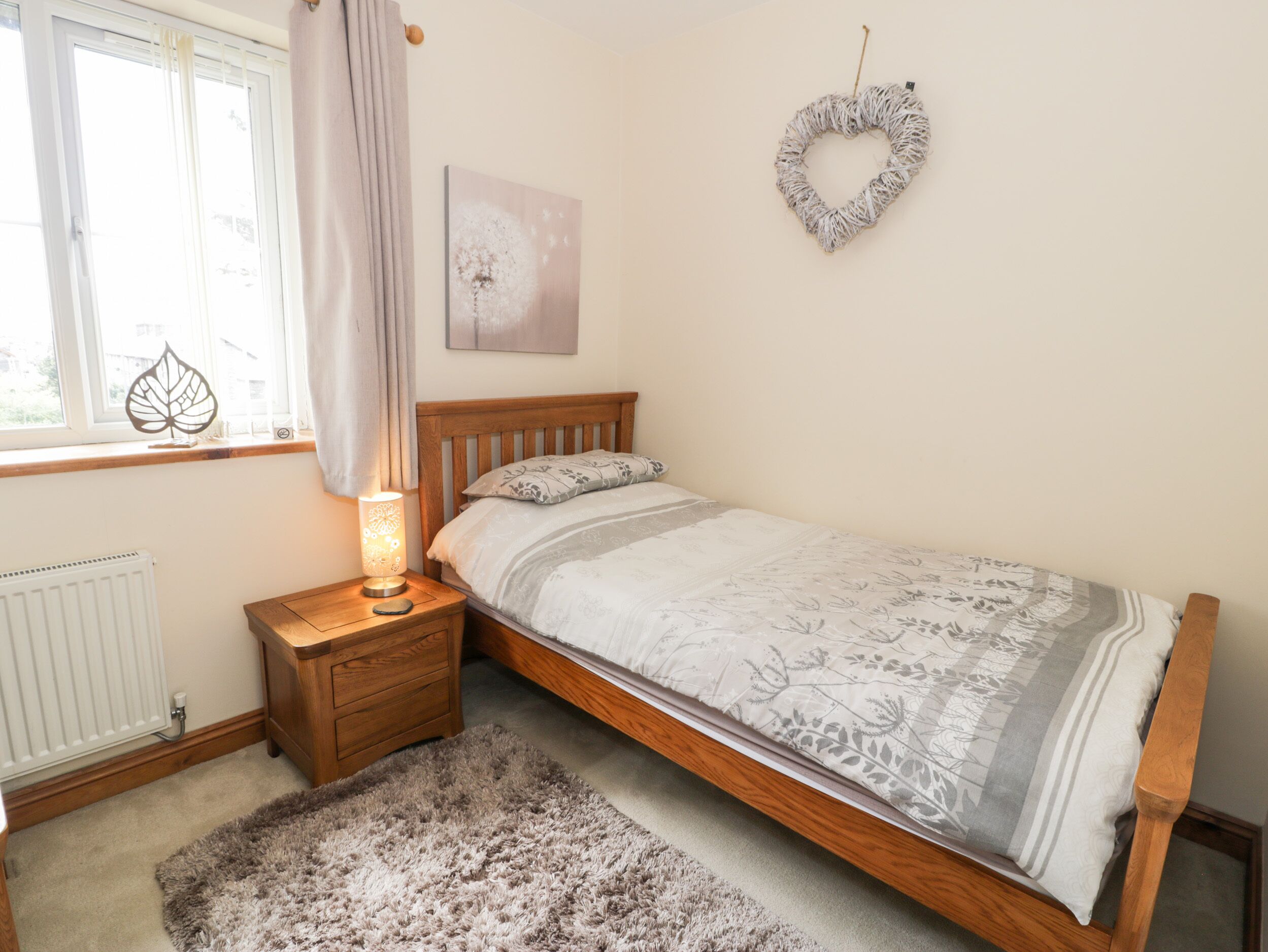 A bedroom with a wooden bed, a nightstand with a lamp, a radiator, a window with curtains, and a wall decoration.