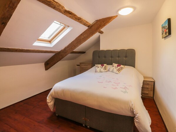 A bedroom with a sloped ceiling, a bed with floral bedding, a nightstand, and a small dresser. - Lower West Curry Farmhouse (Launceston)