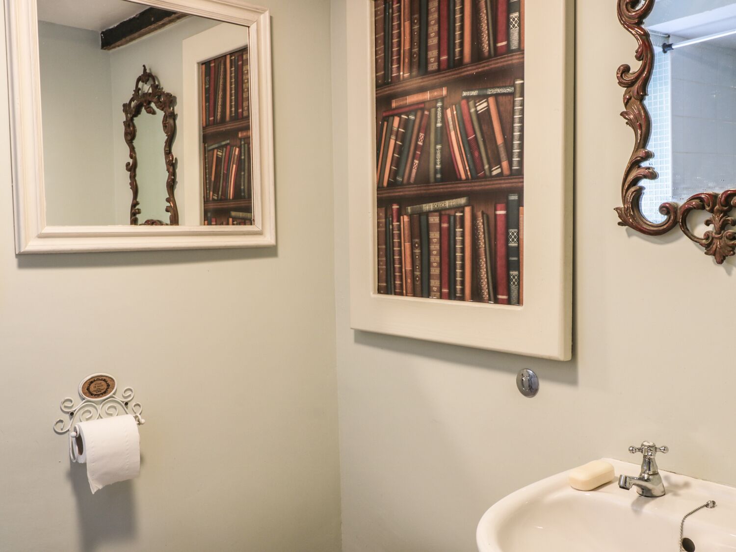 A bathroom with a toilet paper holder, a sink with a soap bar, and a mirror reflecting a bookshelf.