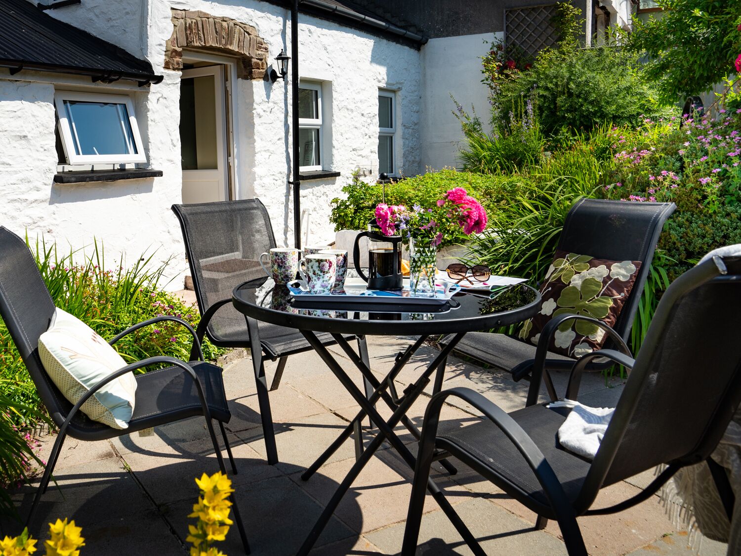 A patio with a round table and four chairs, surrounded by a garden with various plants and flowers.