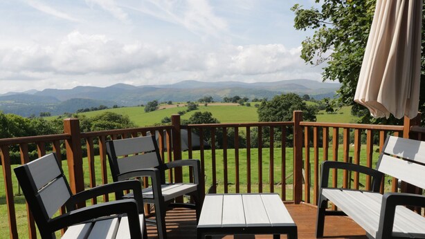 A wooden deck with outdoor furniture, including chairs and a table, overlooking a green landscape with hills in the distance. - Hen Stabl (Llanrwst)