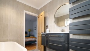 Un baño moderno con una bañera independiente, una puerta de madera que da a un dormitorio y un espejo grande sobre el lavabo.