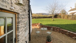 A stone house with a white door, a small patio with a table and chairs, and a grassy field in the background. - Ivy Cottage (Malvern)