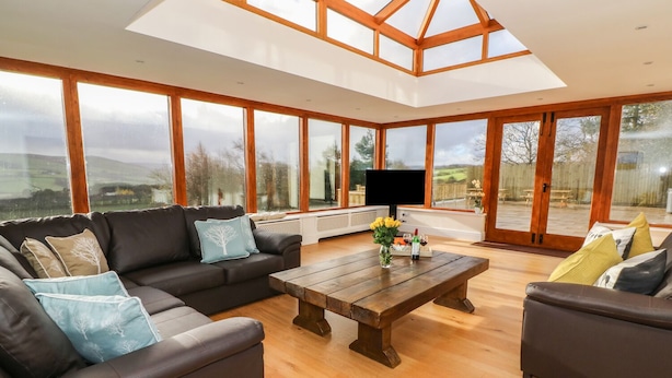 A spacious living room with a large wooden coffee table, two sofas, and a TV. The room has large windows offering a view of the outdoors. - Oakwood House (Rhayader)