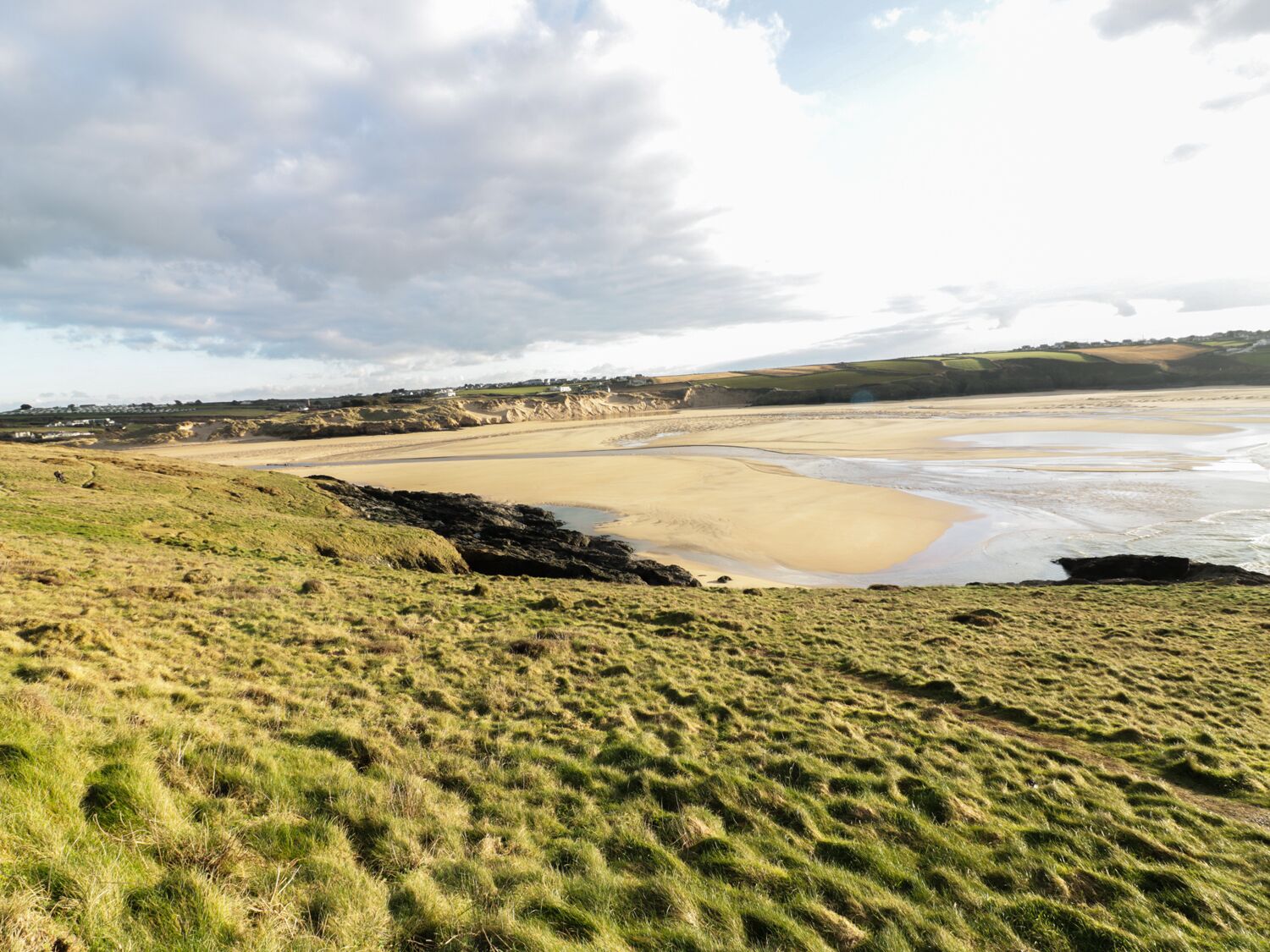 Un paysage côtier avec des champs couverts d’herbe, une plage de sable et un village situé au loin.