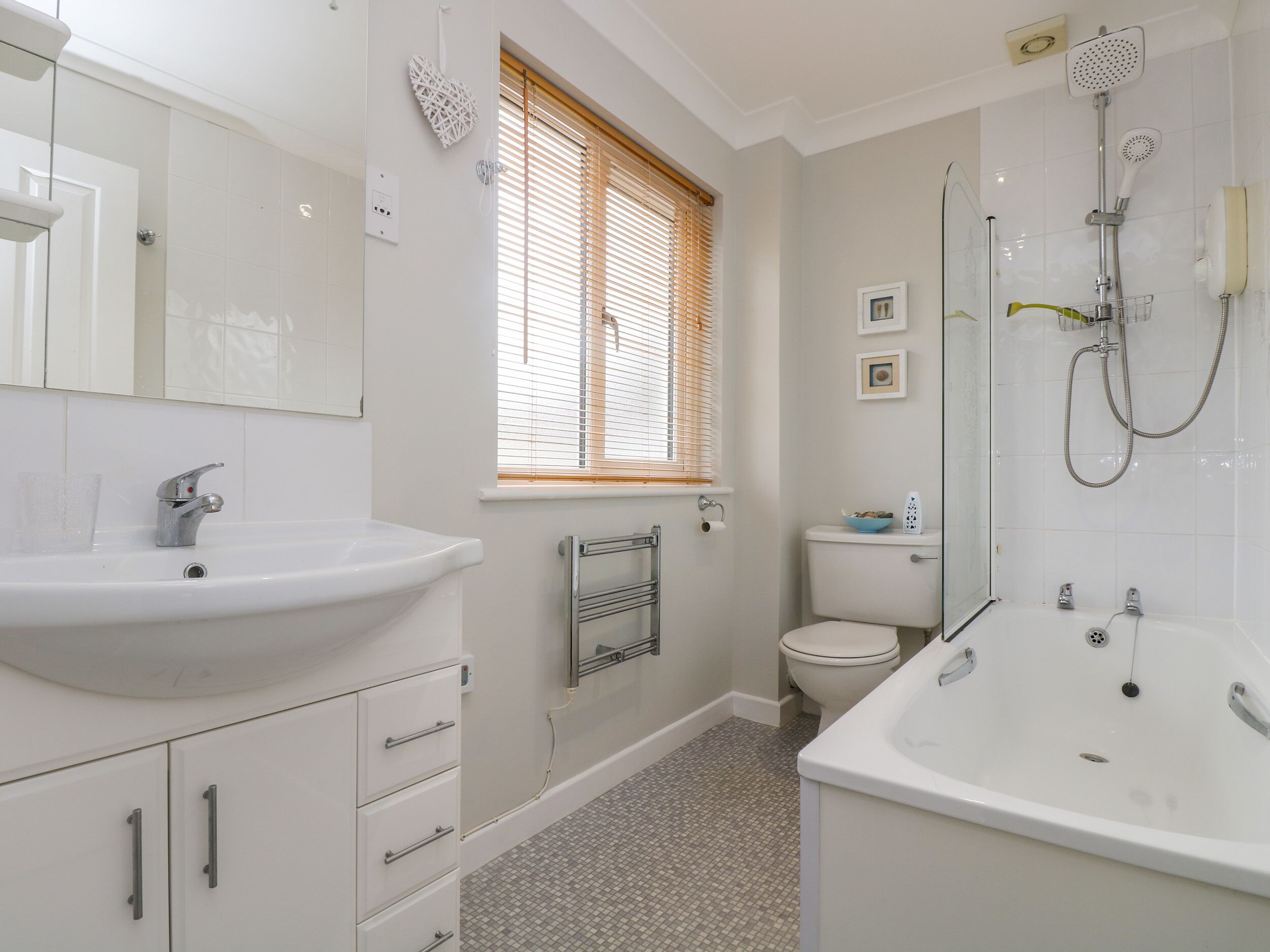 A modern bathroom with a white sink, vanity, and bathtub. There is a window with blinds, a toilet, and a shower with a handheld showerhead.