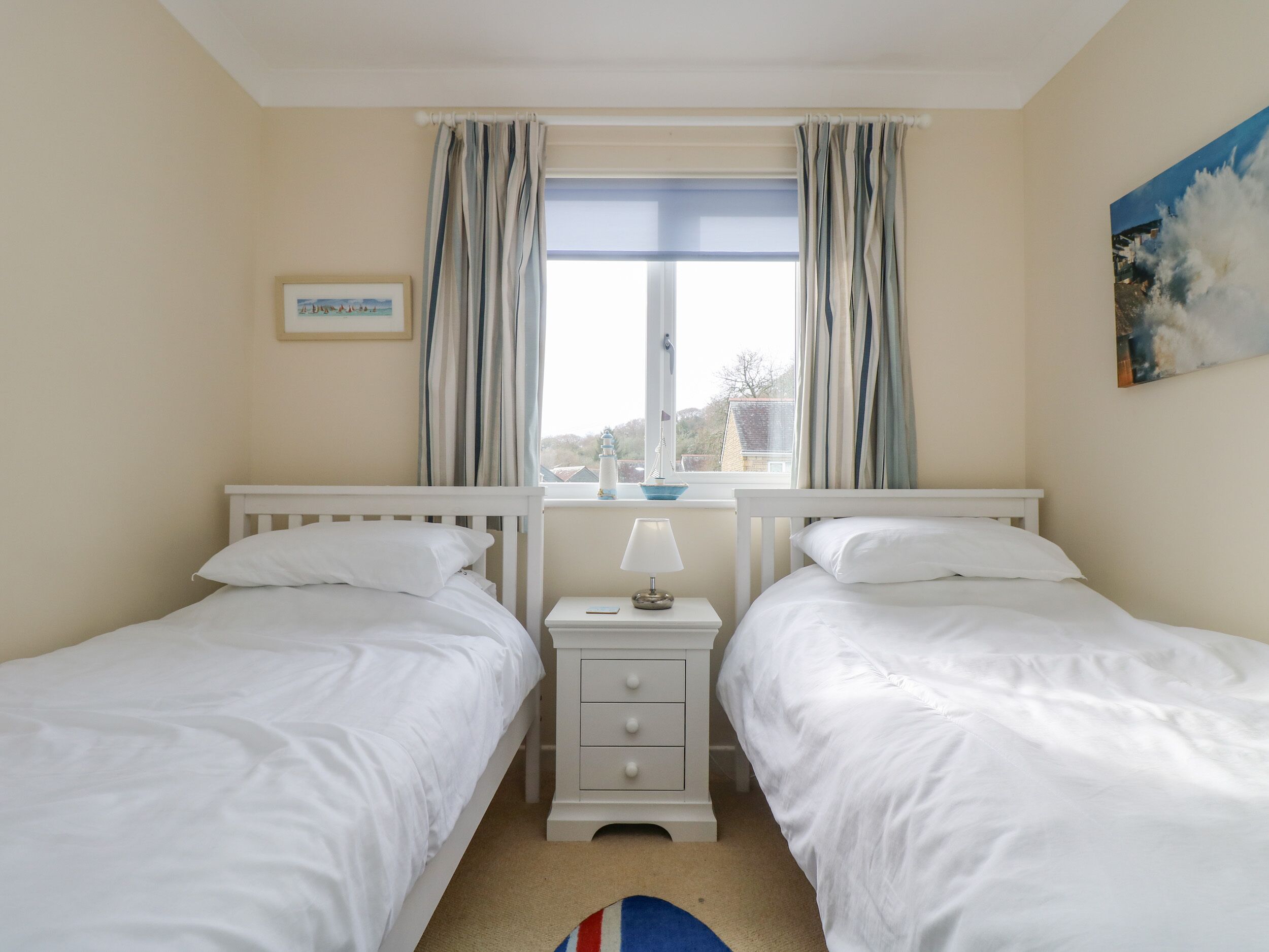 A bedroom with two single beds, a nightstand with a lamp, a framed picture on the wall, and a window with curtains.