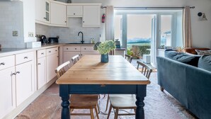 A modern kitchen with white cabinets, a wooden dining table, and a view through the open door. - Stag Cottage (Penrith)