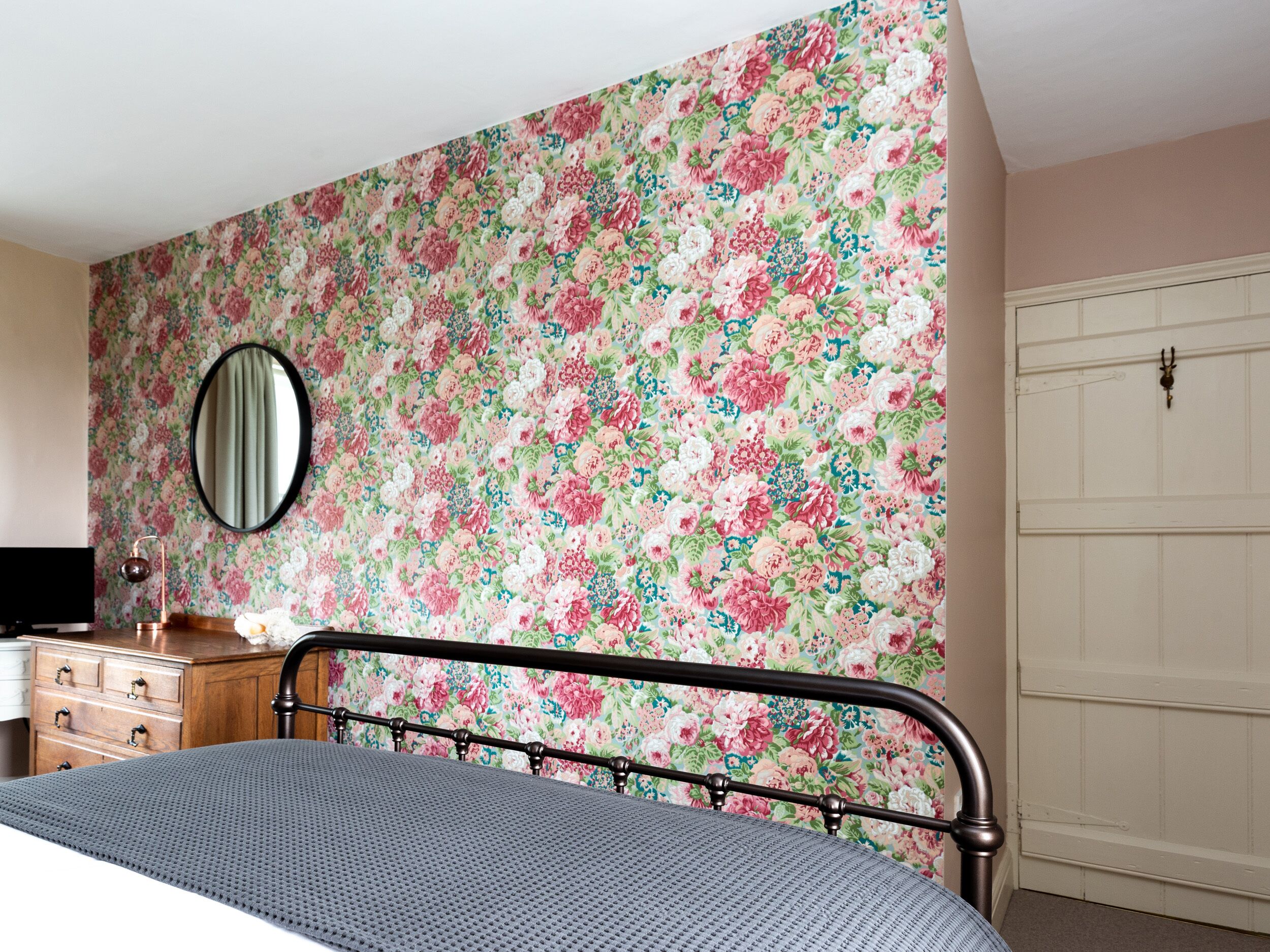 A bedroom with a floral wallpaper, a bed with a metal frame, a dresser, a mirror, and a white door.