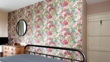 A bedroom with a floral wallpaper, a bed with a metal frame, a dresser, a mirror, and a white door.