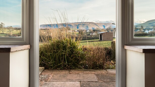 A view through a window of a stone-paved area with grass and plants, leading to a small wooden structure labeled "French Lake." - Stag Cottage (Penrith)