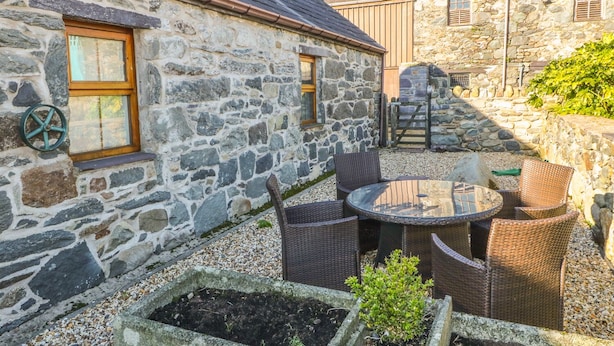 A stone building with a tiled roof, a small outdoor seating area with wicker chairs and a table, and a garden bed with plants. - Beudy Bach (Caernarfon)
