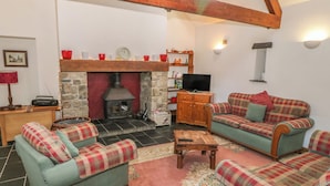 A cozy living room with a stone fireplace, wooden beams, a television, and plaid-patterned furniture. - Beudy Bach (Caernarfon)