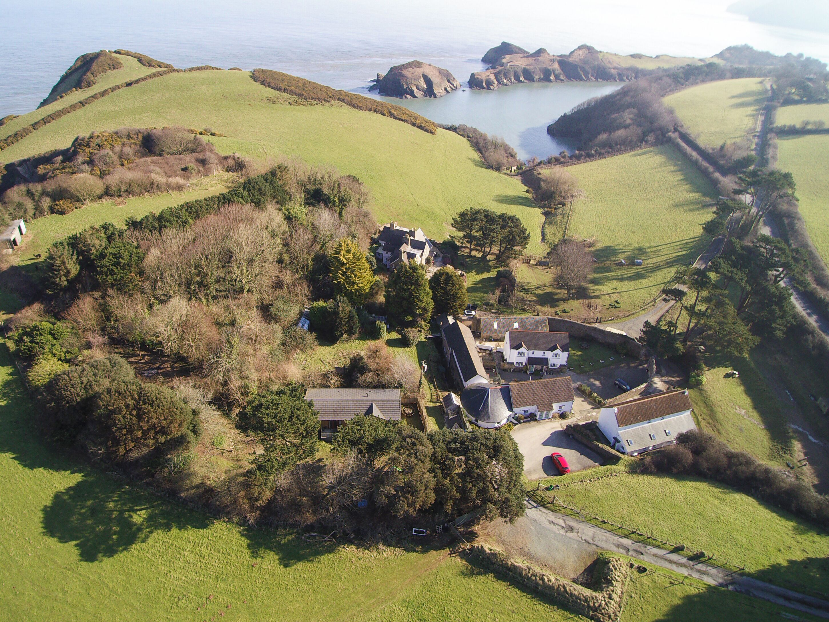 An aerial view of a rural property with multiple buildings, including a large house with a prominent roof, smaller structures, and a red car parked near the main house. The property is surrounded by green fields and a body of water, with hills in the background.