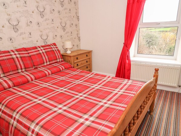 Un dormitorio con colcha escocesa, mesita de noche de madera, lámpara y ventana con cortinas rojas.