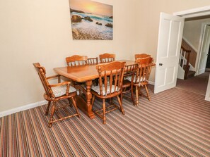 A dining area with a wooden table and chairs, a painting of a beach scene on the wall, and a striped carpet. - The Farmhouse (Llanfairpwllgwyngyll)