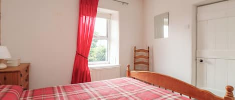 A bedroom with a bed, a wooden chair, a window with red curtains, a bedside table with a lamp, and a white door.