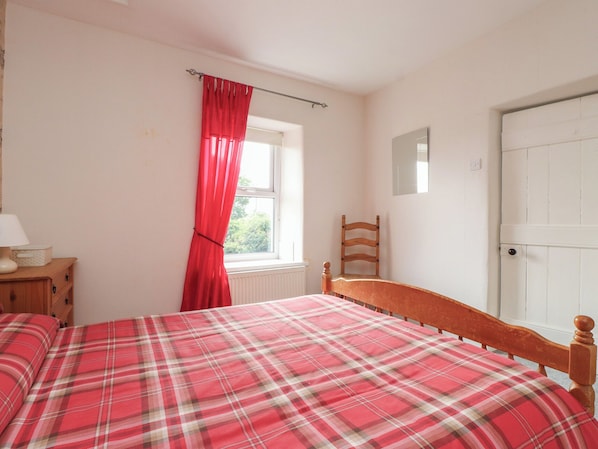 Un dormitorio con cama, una silla de madera, una ventana con cortinas rojas, una mesita de noche con lámpara y una puerta blanca.