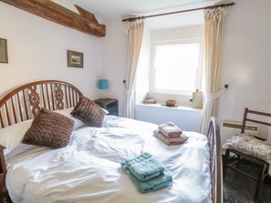 A bedroom with a wooden headboard, a bed with white linens, two pillows, a bedside table with a lamp, a window with curtains, a radiator, and a chair. - Mill Cottage (Kendal)