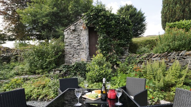 An outdoor dining area with a stone wall, lush greenery, and wicker furniture. - Mill Cottage (Kendal)