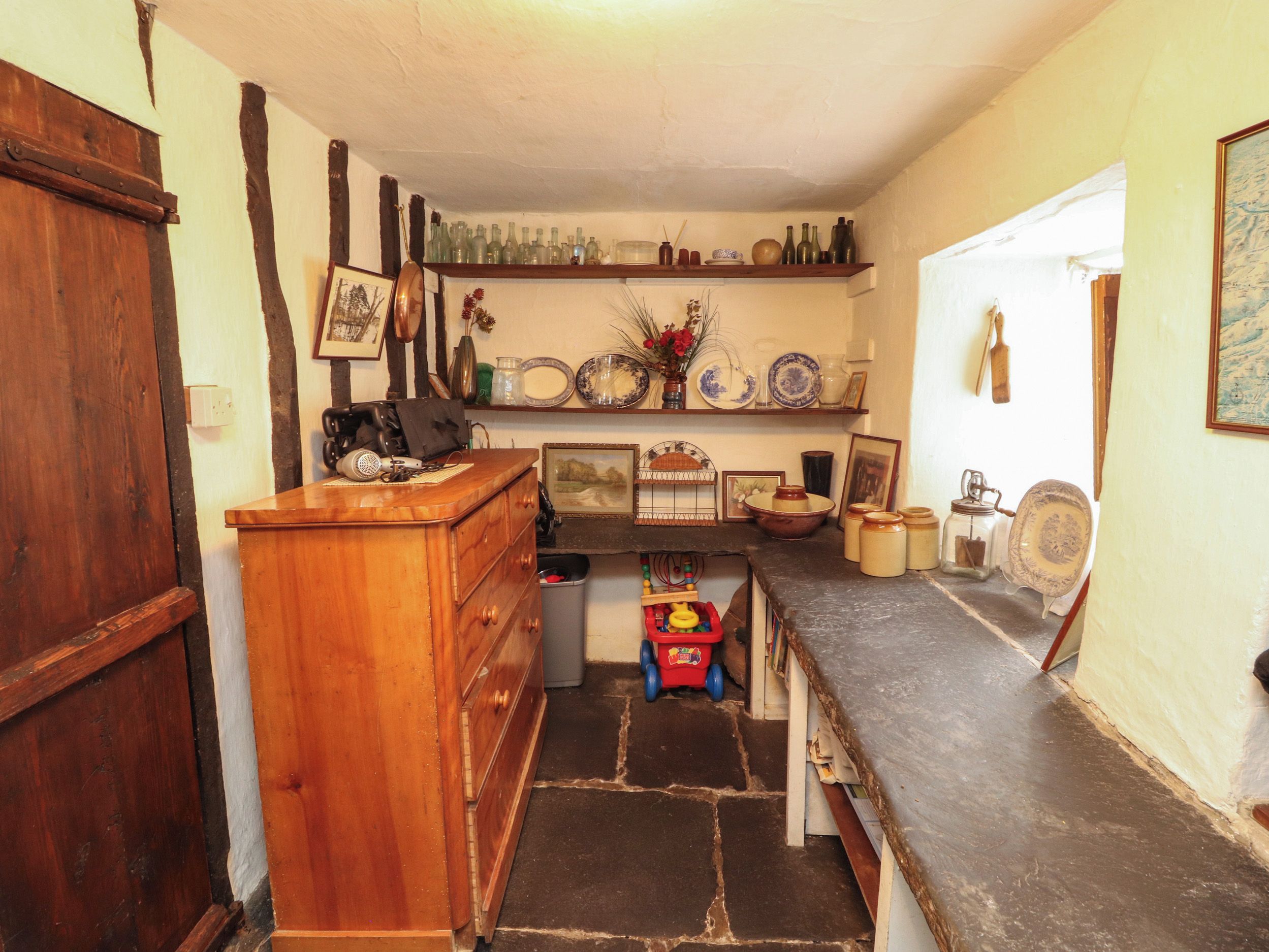 A kitchen with a wooden counter, a sink, and a stove. There are shelves with dishes and a trash bin.