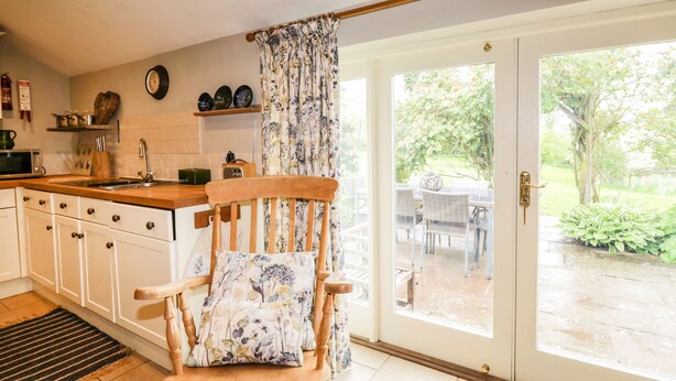 A kitchen with white cabinets, a wooden chair with a floral cushion, a sink, and a window with a view of the garden.
