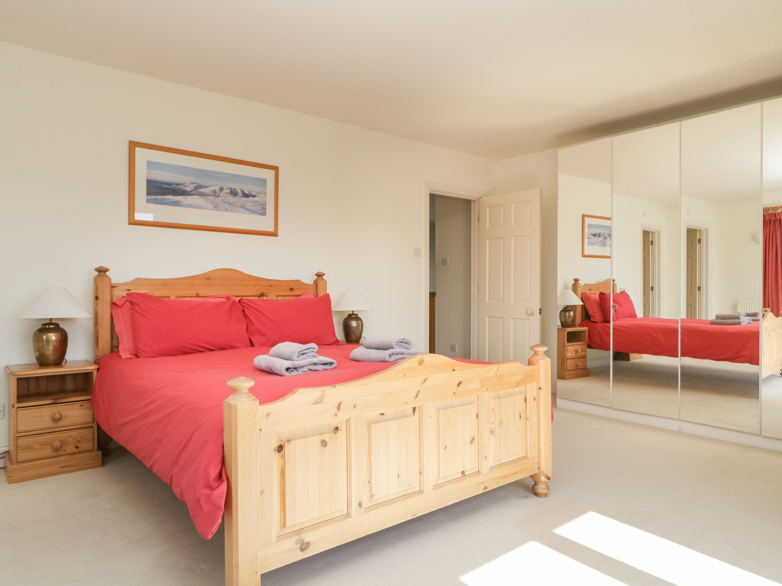 A bedroom with a wooden bed, red bedding, a bedside table with a lamp, a mirror, and a framed picture on the wall.
