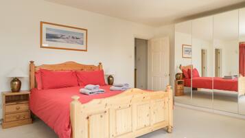 A bedroom with a wooden bed, red bedding, a bedside table with a lamp, a mirror, and a framed picture on the wall.