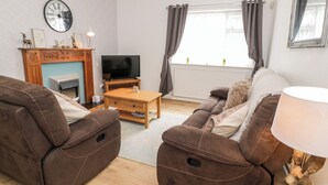A living room with brown leather sofas, a wooden coffee table, a flat-screen TV, a fireplace, and a window with curtains. - Fairways (Prestatyn)