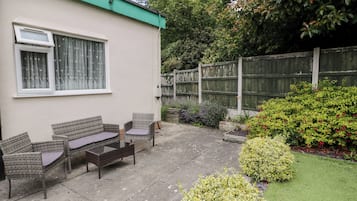 Une terrasse dotée de meubles en osier et d’une petite table, entourée d’un jardin où poussent différentes plantes et délimitée par une clôture en bois.