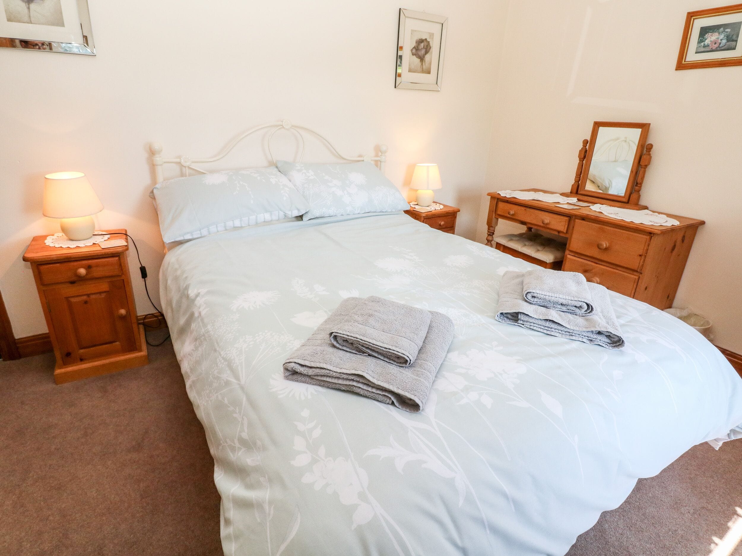 A neatly made bed with floral bedding, two bedside tables with lamps, a wooden nightstand, and a dresser with a mirror.