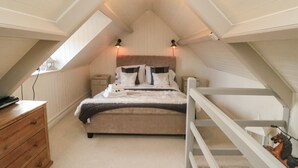 Une chambre avec un plafond en pente, un lit avec des oreillers et une couverture, une table de chevet, une commode et une petite étagère sur laquelle est posé un objet décoratif.