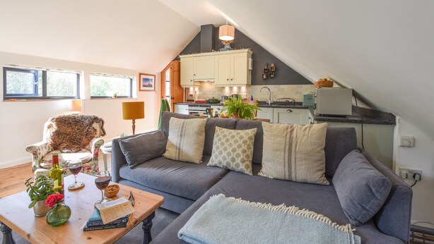 A cozy living room with a grey sofa, a wooden coffee table, and a small dining table. There is a kitchen in the background with white cabinets and a hanging pot. - The Linhay (Okehampton)