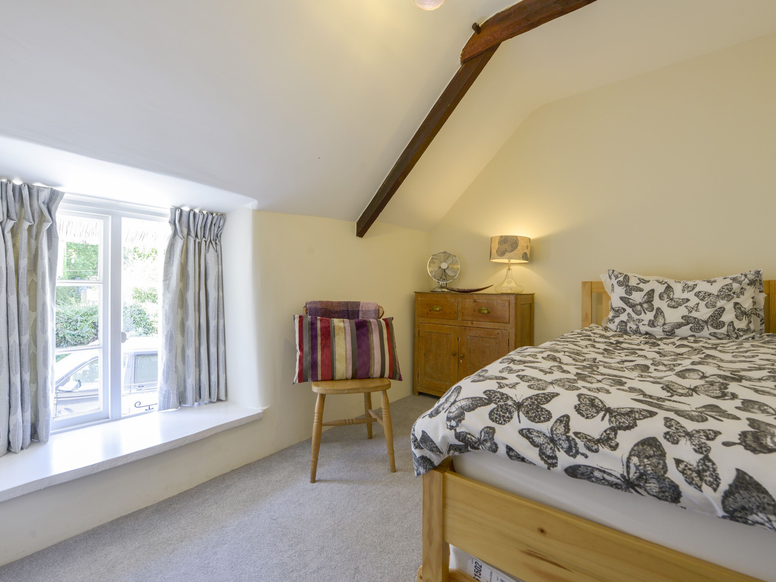 A bedroom with a bed, a wooden dresser, a window with curtains, and a fan on the dresser.