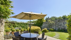 A round table with wicker chairs and a beige umbrella in a garden setting.