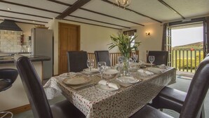 A dining table set for six with a floral tablecloth, surrounded by chairs, in a room with exposed wooden beams and a view of the outdoors.