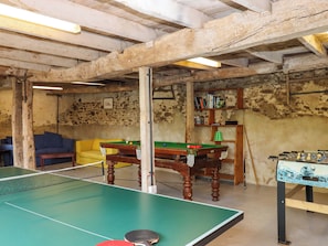 A room with a ping pong table, a pool table, and a foosball table. There is a sofa and a bookshelf in the background.