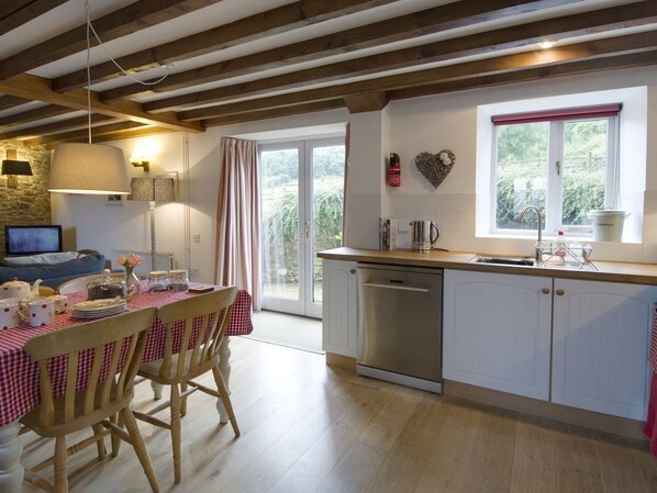 A cozy kitchen with wooden beams, a dining table set with dishes, and a window overlooking greenery. - Whitcombe Cottage (Honiton)