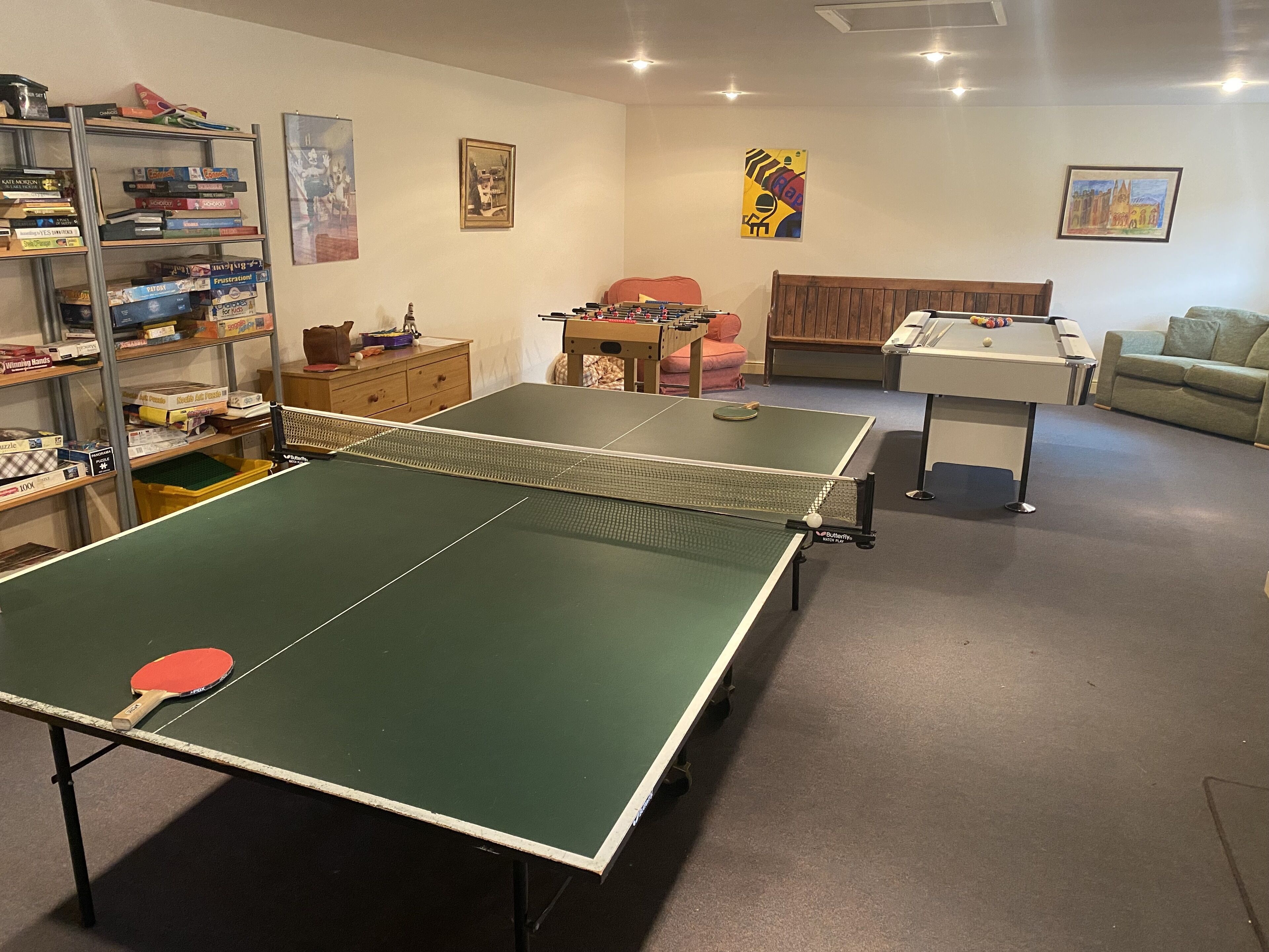 A room with two ping pong tables, a foosball table, and shelves with board games.