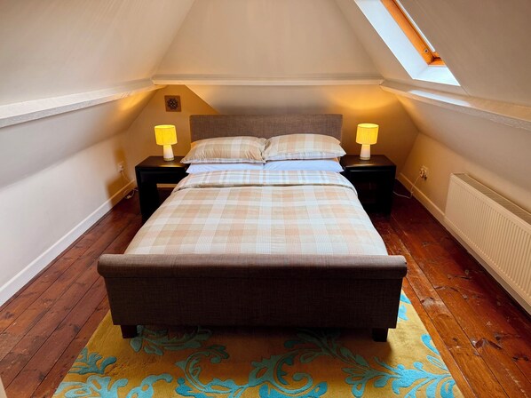 A bedroom with a sloped ceiling, a bed with a headboard, two bedside tables with lamps, a radiator, and a patterned rug. - Honey House (Okehampton)