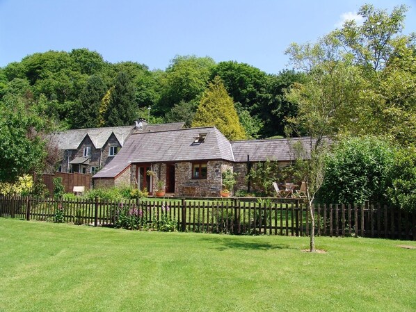 A stone house with a sloped roof, a fenced yard, and lush greenery in the background.