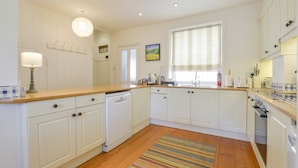 A kitchen with white cabinets, a wooden countertop, a dishwasher, a window with blinds, and a striped rug on the floor. - 4 Coastguards Cottage (Kingsbridge)
