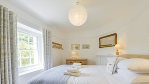 A neatly made bed with a blue blanket, a bedside table with a lamp, and a window with curtains. - 4 Coastguards Cottage (Kingsbridge)