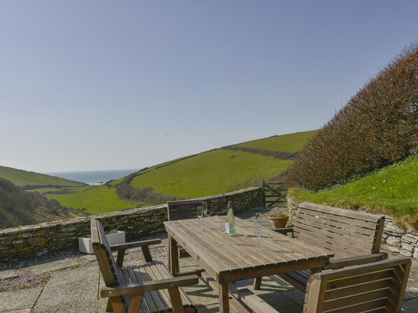 A wooden table and benches set against a stone wall with a view of rolling hills and the sea.
